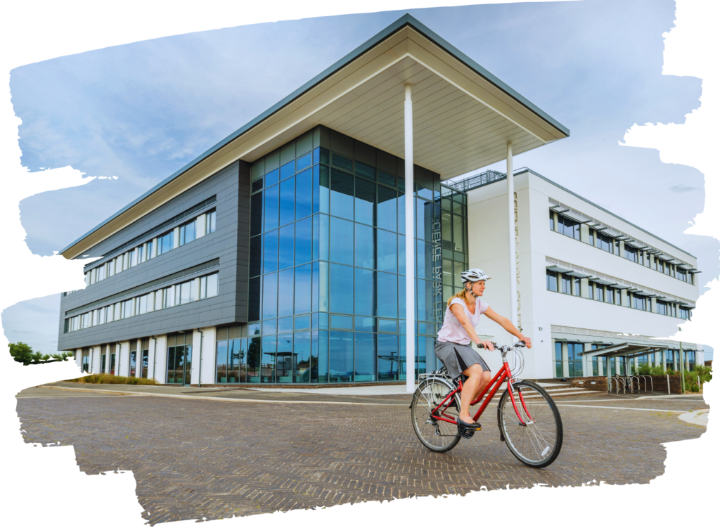 The main building at Exeter Science Park, with a woman on a red bike cycling past. Modern builidng, showing lots of grey cladding and a tall glass atrium, with a flat roof that over hangs the atrium in a dramatic manner.