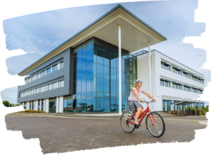 The main building at Exeter Science Park, with a woman on a red bike cycling past. Modern builidng, showing lots of grey cladding and a tall glass atrium, with a flat roof that over hangs the atrium in a dramatic manner.