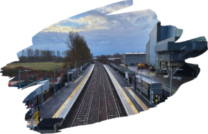 View south over the rail tracks as they pass through Marsh Barton rail station. Green fields to left, and side aspect of an industrial building, which is the Exeter energy from waste plant.