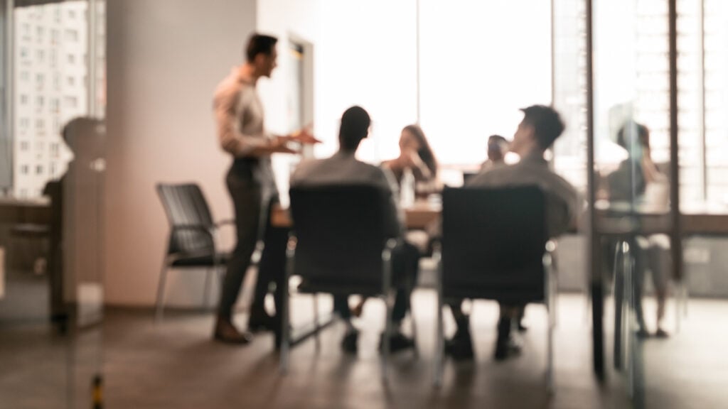 A group of individuals in a meeting room. One person stands at the forefront, appearing to address the others who are seated around a table. The setting suggests a professional environment, possibly an office with large windows that allow natural light to fill the room. The focus is not on individual details but rather on the collective scene of a workplace meeting in progress.